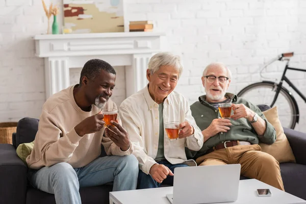 Sonriendo amigos interracial utilizando el ordenador portátil y la celebración de té en casa - foto de stock