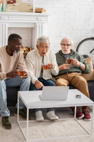Sorprendidos hombres multiétnicos sosteniendo té y mirando a la computadora portátil en casa - foto de stock