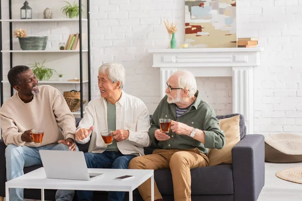 Sonriente asiático hombre con té mostrando como cerca multiétnico amigos y dispositivos en casa - foto de stock