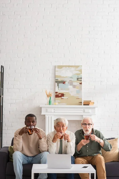 Amigos multiculturales impactados sosteniendo té cerca de gadgets en casa - foto de stock