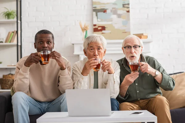 Schockierte multiethnische Freunde halten Tassen in der Nähe von Geräten im Wohnzimmer — Stockfoto