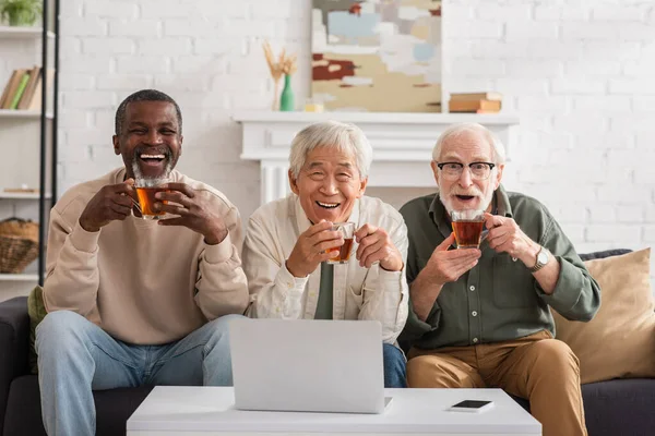 Heureux retraités multiethniques tenant des tasses de thé près d'un ordinateur portable et d'un téléphone portable à la maison — Photo de stock