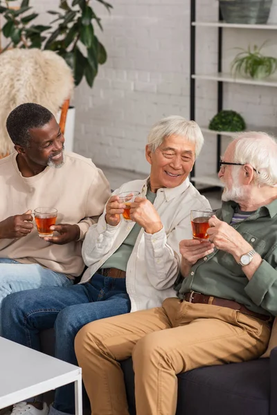 Amigos interracial positivos sosteniendo té y hablando en el sofá en casa - foto de stock