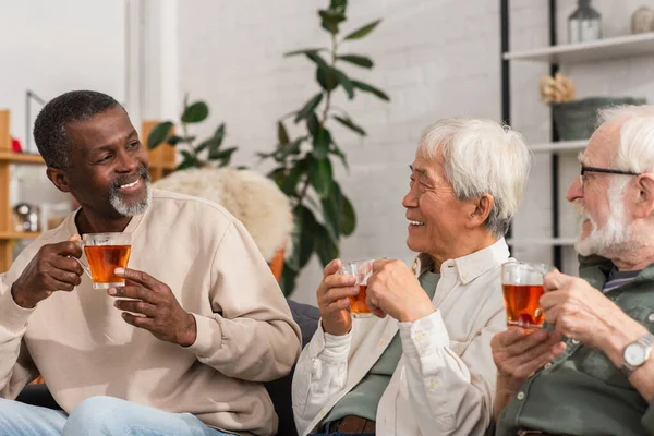 Positivo afroamericano hombre sosteniendo té cerca de amigos multiétnicos en casa - foto de stock