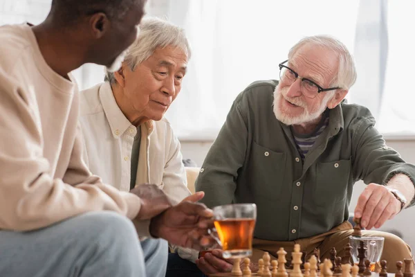 Sorrindo homem jogando xadrez com amigos inter-raciais perto de chá em casa — Fotografia de Stock