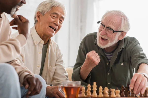 Eccitato uomo anziano che gioca a scacchi con amici multietnici a casa — Foto stock