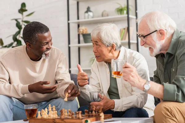 Hombre asiático sosteniendo té y mostrando como signo cerca de amigos multiétnicos y ajedrez en casa - foto de stock