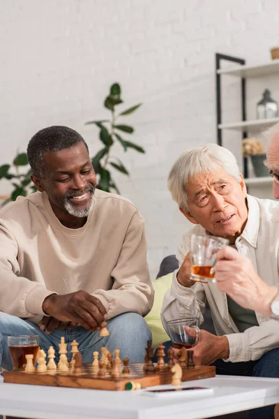 Multiethnic senior friends with tea playing chess near blurred smartphone at home — Stock Photo