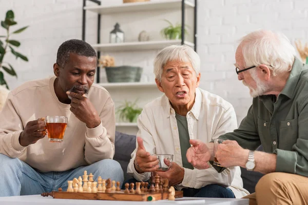 Interracial elderly friends with tea pointing at chess at home — Stock Photo