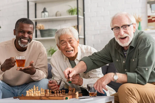 Alegre multiétnicos viejos amigos mirando a la cámara cerca de ajedrez y té en casa - foto de stock