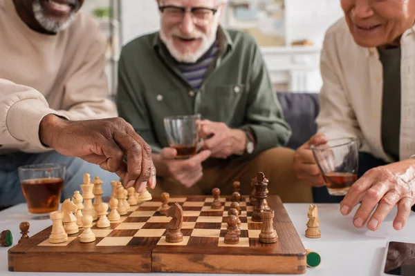 Hombre afroamericano jugando ajedrez cerca de teléfonos inteligentes, té y amigos en casa - foto de stock