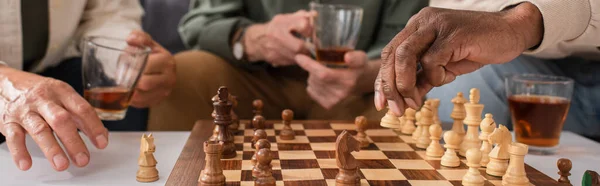 Vista recortada de amigos multiétnicos con té jugando ajedrez en casa, pancarta - foto de stock