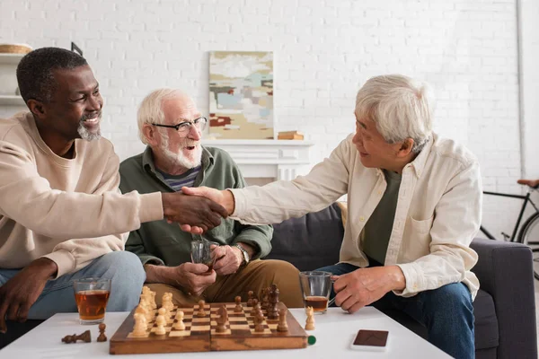 Amigos multiétnicos estrechando manos cerca del té y el ajedrez en casa - foto de stock