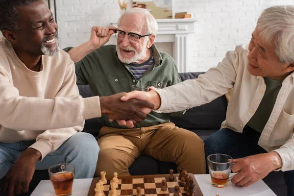 Homme âgé souriant tenant le thé près d'amis interraciaux serrant la main et les échecs à la maison — Photo de stock