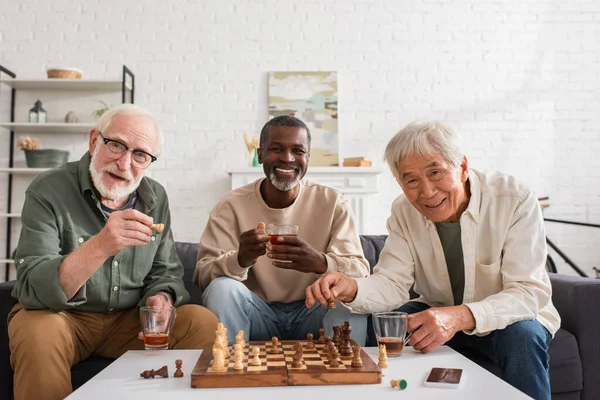 Cheerful interracial friends holding tea and chess figures at home — Stock Photo