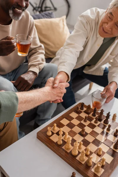 High angle view of interracial friends shaking hands near african american man with tea and chessboard — Stock Photo