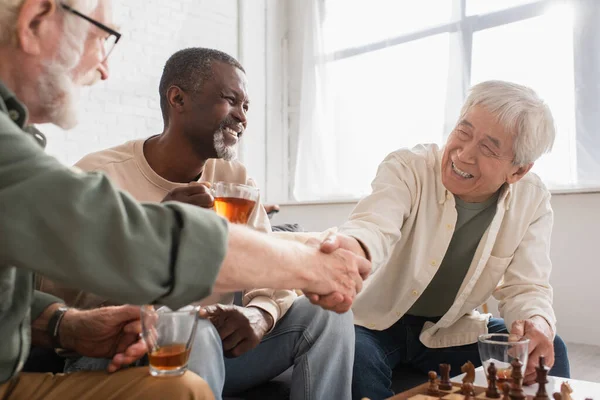 Interracial pensioners shaking hands near chessboard and african american friend with tea at home — Stock Photo