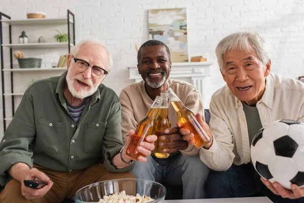 Позитивні міжрасові старші друзі з футболом і пивом дивляться на камеру вдома — стокове фото