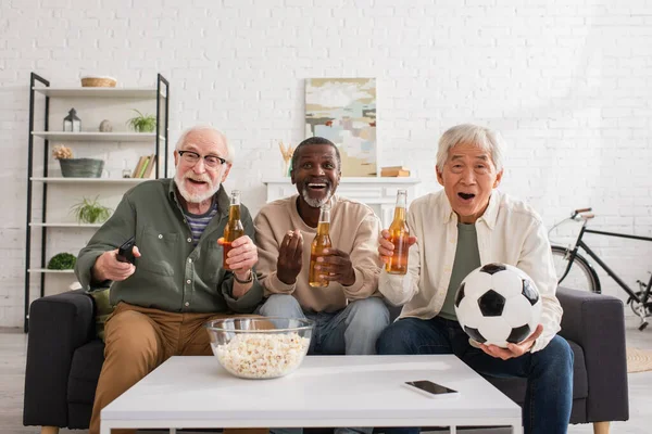 Amici anziani multiculturali con calcio e birra guardando la tv a casa — Foto stock