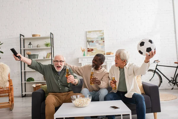 Amigos Interraciales sosteniendo fútbol y cerveza cerca del hombre con mando a distancia en casa - foto de stock