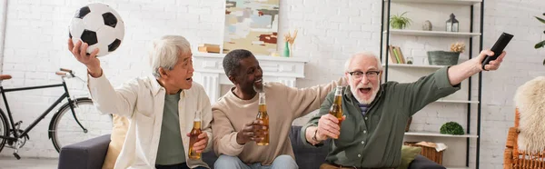 Uomo felice che tiene telecomando vicino agli amici multietnici con birra e calcio a casa, banner — Foto stock