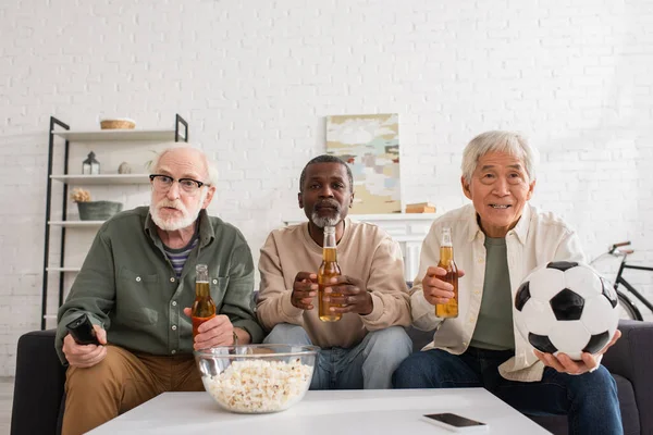 Amigos multiétnicos mayores sosteniendo cerveza y fútbol cerca de palomitas de maíz en casa - foto de stock