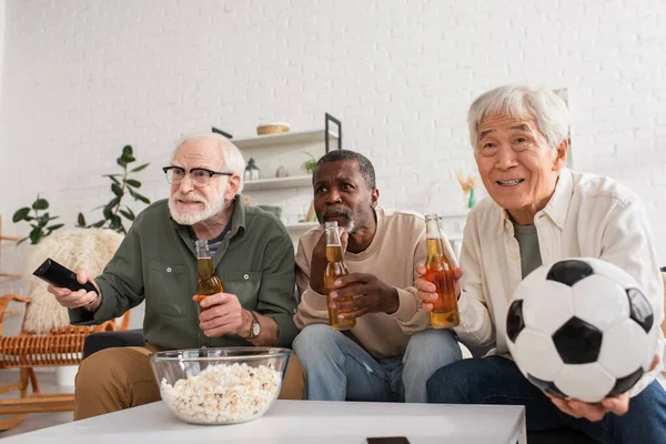 Estressado interracial amigos segurando cerveja e futebol perto de pipocas em casa — Fotografia de Stock