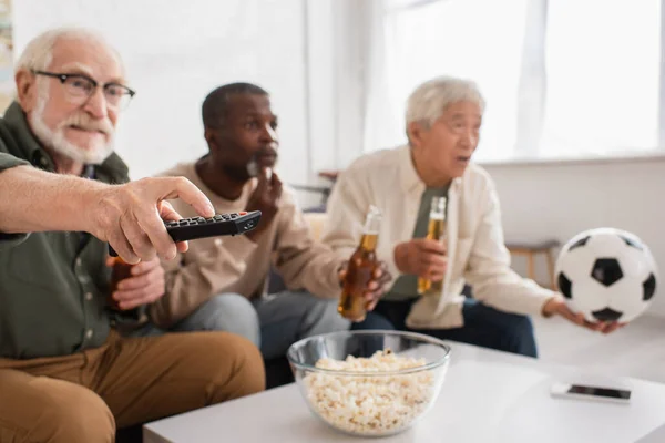 Hombre mayor sosteniendo mando a distancia cerca borrosa interracial amigos con cerveza en casa - foto de stock