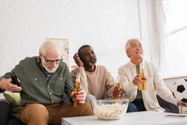 Stressed interracial pensioners holding beer and football near popcorn in living room — Stock Photo