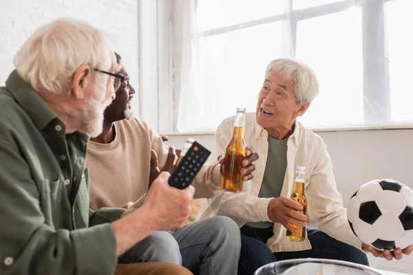 Asiatico uomo holding calcio vicino interrazziale amici con birra a casa — Foto stock