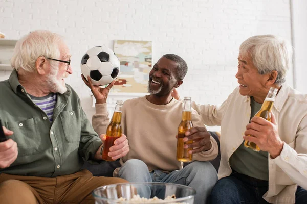 Alegre multicultural homens segurando cerveja e futebol em casa — Fotografia de Stock