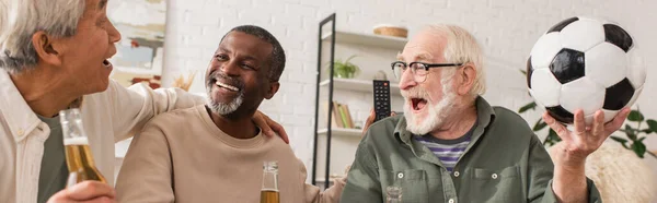 Hombres mayores interraciales sosteniendo botellas de cerveza y fútbol en casa, pancarta - foto de stock