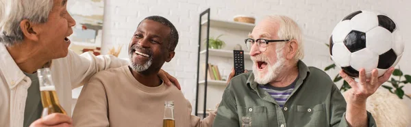 Interracial senior men holding beer bottles and football at home, banner — Stock Photo