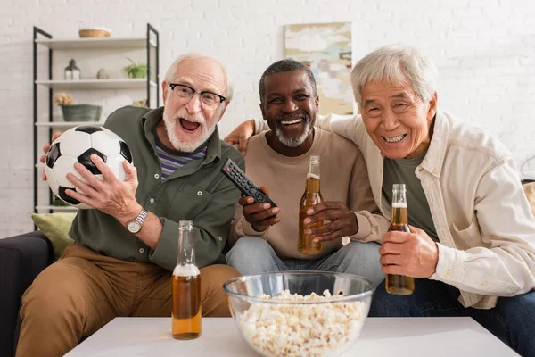 Uomini multietnici positivi che tengono bottiglie di birra e calcio sul divano a casa — Foto stock