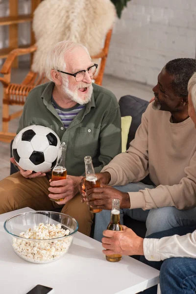 Sorridente interrazziale uomini con birra guardando amico in possesso di calcio a casa — Foto stock