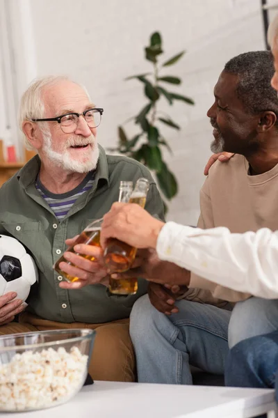 Ancianos hombres multiétnicos tintineo cerveza con amigo celebración de fútbol en casa - foto de stock