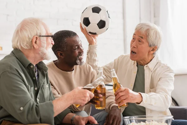 Asiático homem segurando futebol perto de amigos multiétnicos com cerveja em casa — Fotografia de Stock