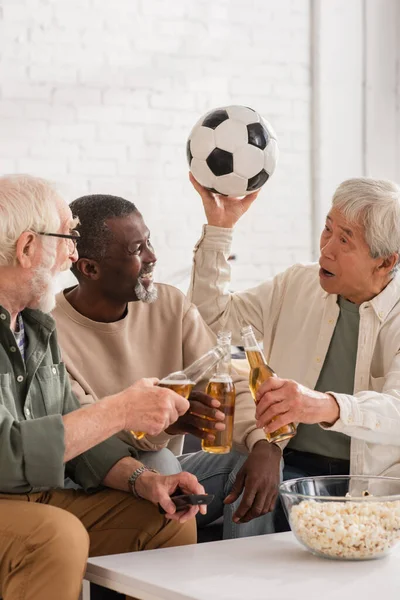 Positive multiethnic men holding beer and football near popcorn at home — Stock Photo