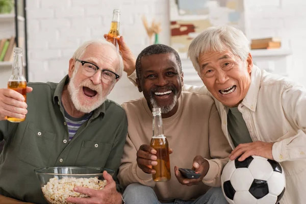 Sorridenti amici anziani interrazziale con birra, popcorn e telecomando guardando la fotocamera a casa — Foto stock