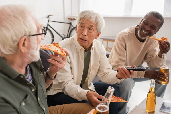 Asiático homem segurando controle remoto perto de amigos inter-raciais com cerveja e pizza em casa — Fotografia de Stock