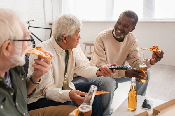 Sonriente hombre afroamericano sosteniendo cerveza y pizza cerca de amigos multiétnicos con controlador remoto en casa - foto de stock