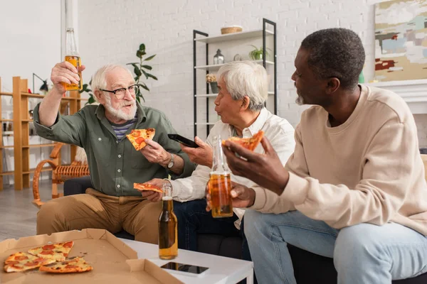 Uomo anziano che tiene pizza e birra vicino agli amici multietnici con telecomando a casa — Foto stock