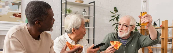 Старший мужчина держит пиццу и пиво возле мультикультурных друзей с пультом дистанционного управления на дому, баннер — стоковое фото