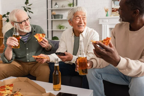 Älterer asiatischer Mann hält Fernbedienung in der Hand und spricht mit multiethnischen Freunden bei Bier und Pizza zu Hause — Stockfoto