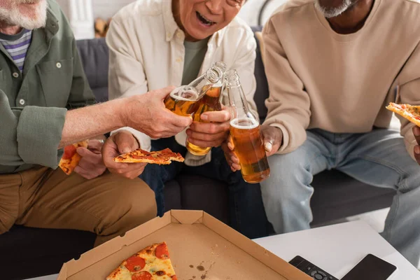 Vista cortada de pensionistas inter-raciais clinking com cerveja e segurando pizza em casa — Fotografia de Stock