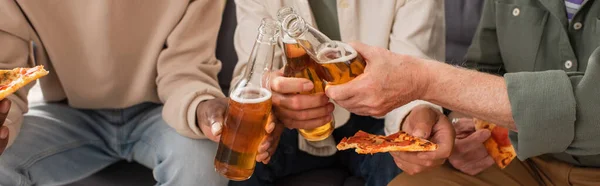 Vista recortada de amigos mayores multiétnicos sacudiendo la cerveza cerca de la pizza en casa, pancarta - foto de stock