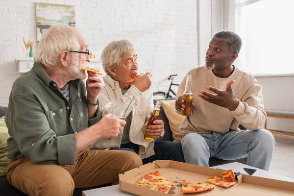 Африканський американець з пивом розмовляє зі старшими міжрасовими друзями, які їдять піцу вдома. — стокове фото