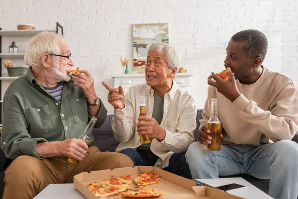 Старший азиат держит пиво и указывает пальцем на межрасовых друзей, поедающих пиццу дома — стоковое фото