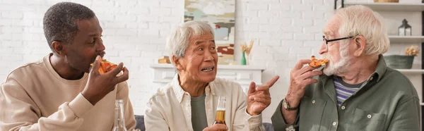 Anziani asiatico uomo holding birra e puntare con dito vicino multietnico amici mangiare pizza a casa, striscione — Foto stock
