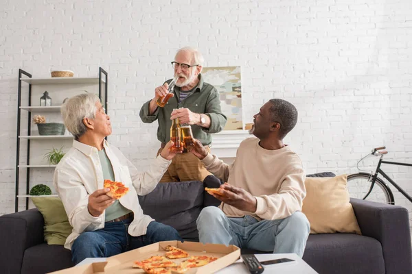 Ältere multiethnische Freunde, die zu Hause neben Pizza und Fernbedienung Bier trinken — Stockfoto